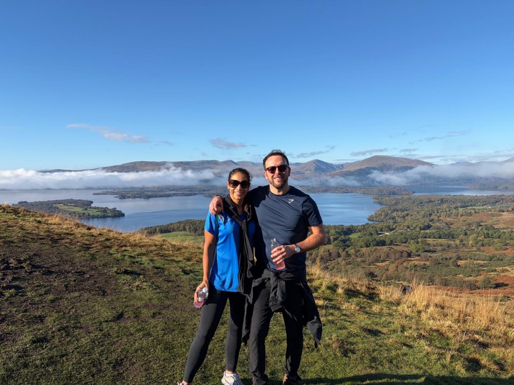 A couple standing on a hillside overlooking a scenic lake and mountainous landscape on a clear day.